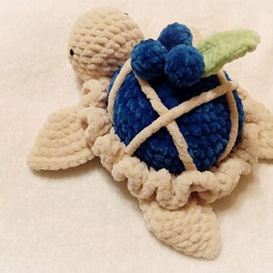 May include: A crocheted stuffed turtle toy with a blue shell and a green leaf on top. The shell is decorated with white yarn and has a cluster of blue yarn balls on top.