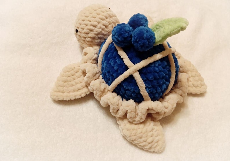 May include: A crocheted stuffed turtle toy with a blue shell and a green leaf on top. The shell is decorated with white yarn and has a cluster of blue yarn balls on top.