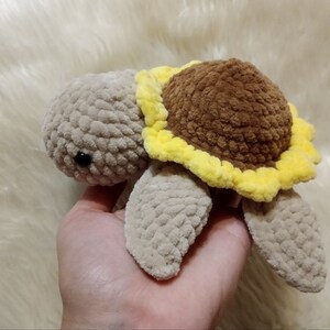 May include: A crocheted stuffed turtle toy with a brown sunflower on its back. The turtle is beige and the sunflower has yellow petals.