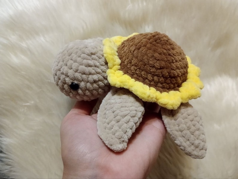 May include: A crocheted stuffed turtle toy with a brown sunflower on its back. The turtle is beige and the sunflower has yellow petals.