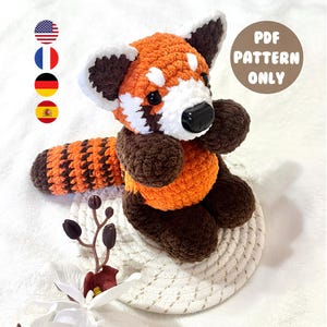 Op de afbeelding: Een gehaakte rode panda knuffel. De knuffel is gemaakt van oranje, bruin en wit garen, met een zwarte neus en kleine zwarte ogen. De tekst "PDF PATTERN ONLY" staat in een bruine cirkel. De knuffel zit op een witte onderzetter.