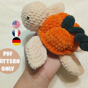 May include: A crocheted turtle amigurumi toy with a beige body, orange shell, and black accents. The toy is being held in a hand.