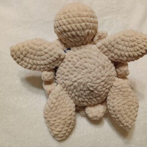 May include: A crocheted plush toy shaped like a turtle. The turtle is beige and has a textured surface. The turtle is lying on its back with its legs and head visible.