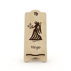 May include: Wooden phone stand with a Virgo zodiac sign and the symbol for Virgo. The stand is light brown and has a cutout for a phone.