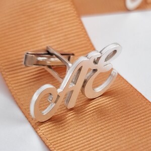 Pode incluir: Um par de botões de punho de prata com as letras "AE" em uma fonte cursiva. Os botões de punho estão em uma fita laranja clara.