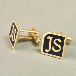 May include: A pair of gold-coloured cufflinks with a black square face. Each cufflink has the gold letters "JS". The cufflinks are displayed against a neutral background.