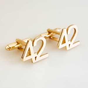 May include: Gold-colored cufflinks with the number 42 in a bold, sans-serif font. The cufflinks are displayed on a white surface, showcasing their polished finish and simple design. The cufflinks are a classic accessory.