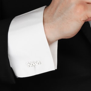 May include: A man wearing a black suit jacket with white dress shirt and cufflinks that say "Kevin" on them.