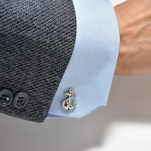 Può includere: Un paio di gemelli d'argento con un design a forma di ancora. I gemelli sono indossati su una camicia azzurra con un blazer blu scuro.