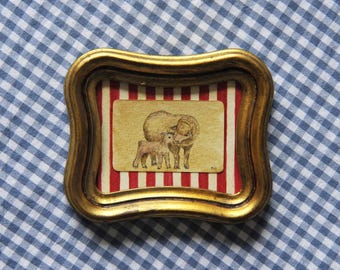 May include: A small, framed artwork with a gold-colored, ornate frame. The artwork features a watercolor illustration of a sheep and a lamb on a red and white striped background. The frame is set against a blue and white checkered fabric.