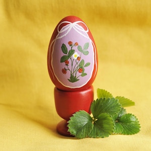 Könnte beinhalten: Ein rotes Holzei auf einem passenden Ständer, verziert mit einem Erdbeerpflanzen-Design in einem lila Oval. Grüne Erdbeerblätter liegen neben dem Ei. Der Hintergrund ist ein gelber Stoff.