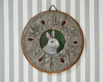 May include: A round, silver-toned metal wall plaque with a raised design of leaves and a small, framed image of a white rabbit. The plaque has a dark wood frame and red glass accents.