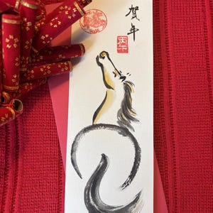 K&ouml;nnte beinhalten: Ein Aquarellgem&auml;lde eines Pferdes in einem kreisf&ouml;rmigen Design mit japanischen Schriftzeichen und einem roten Stempel. Das Kunstwerk befindet sich auf einer wei&szlig;en Karte mit roten Akzenten und dekorativen roten Zylindern mit goldenen Akzenten.
