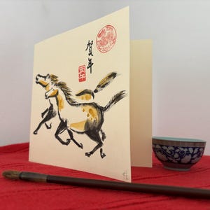 May include: A greeting card with a watercolor painting of two horses in black, yellow, and white. The card has Japanese calligraphy and a red seal. A small blue and white bowl and a calligraphy brush are in the foreground.