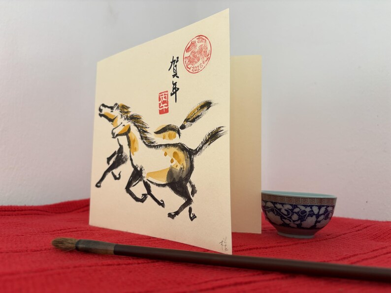 May include: A greeting card with a watercolor painting of two horses in black, yellow, and white. The card has Japanese calligraphy and a red seal. A small blue and white bowl and a calligraphy brush are in the foreground.