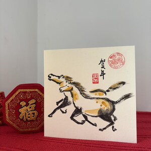 May include: A square greeting card featuring a watercolor painting of two running horses in black, brown, and gold. The card has Japanese text and a red seal stamp. A red box with gold accents is visible.