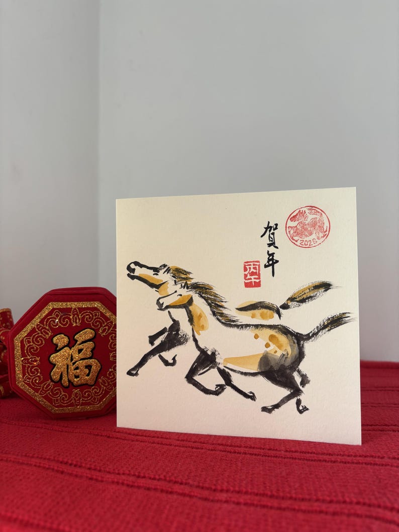 May include: A square greeting card featuring a watercolor painting of two running horses in black, brown, and gold. The card has Japanese text and a red seal stamp. A red box with gold accents is visible.