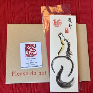 K&ouml;nnte beinhalten: Ein handgemalter Kunstdruck mit einer Pferdeillustration in Schwarz, Wei&szlig; und Gold. Dazu geh&ouml;ren ein Kraftpapierumschlag und ein K&uuml;nstleretikett. Der Hintergrund ist ein roter Stoff. Japanische Schriftzeichen sind enthalten.