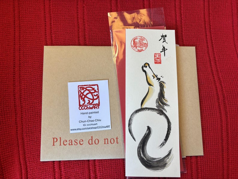 K&ouml;nnte beinhalten: Ein handgemalter Kunstdruck mit einer Pferdeillustration in Schwarz, Wei&szlig; und Gold. Dazu geh&ouml;ren ein Kraftpapierumschlag und ein K&uuml;nstleretikett. Der Hintergrund ist ein roter Stoff. Japanische Schriftzeichen sind enthalten.