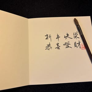 Pode incluir: Cart&atilde;o de felicita&ccedil;&otilde;es aberto com caligrafia chinesa preta e um carimbo vermelho. Um pincel de caligrafia est&aacute; sobre o cart&atilde;o. O fundo &eacute; preto. O cart&atilde;o &eacute; creme.