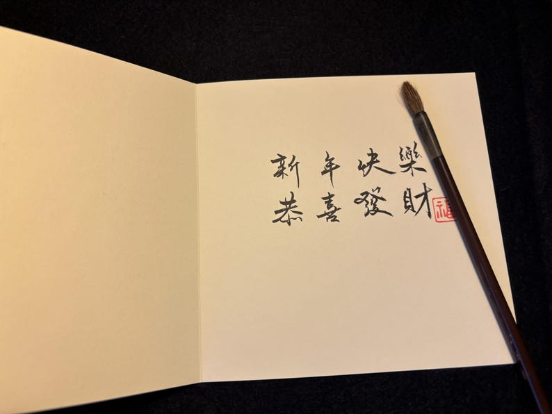 Pode incluir: Cart&atilde;o de felicita&ccedil;&otilde;es aberto com caligrafia chinesa preta e um carimbo vermelho. Um pincel de caligrafia est&aacute; sobre o cart&atilde;o. O fundo &eacute; preto. O cart&atilde;o &eacute; creme.