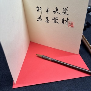 May include: A greeting card with Chinese calligraphy in black ink, featuring the phrases "Happy New Year" and "Wishing you prosperity." The card is open, revealing a red interior and a calligraphy brush.