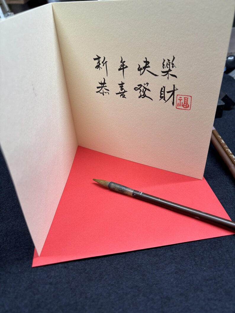 May include: A greeting card with Chinese calligraphy in black ink, featuring the phrases "Happy New Year" and "Wishing you prosperity." The card is open, revealing a red interior and a calligraphy brush.