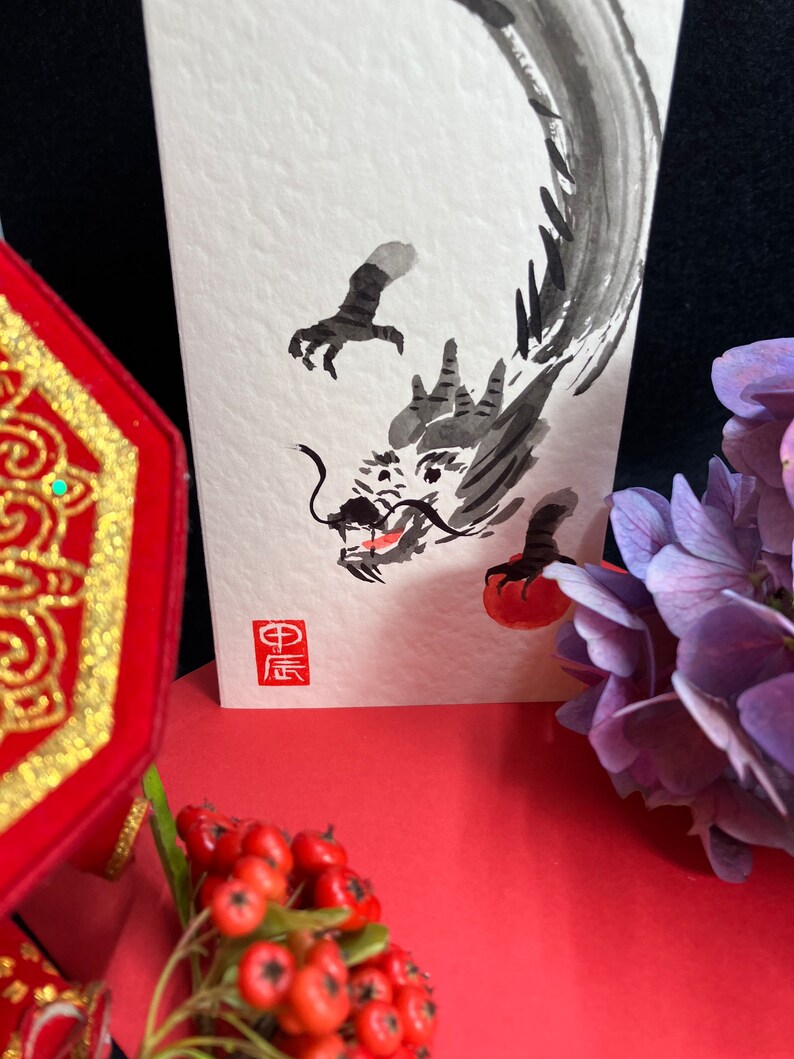Puede incluir: Una pintura de tinta negra de un drag&oacute;n en papel blanco con un c&iacute;rculo rojo y el car&aacute;cter chino para "drag&oacute;n" en tinta roja.