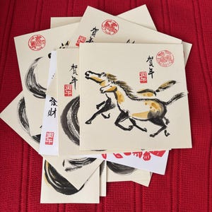 May include: A collection of square greeting cards with Japanese calligraphy and illustrations. The cards feature a running horse, brushstrokes, and red stamps. The background is a red textured surface.