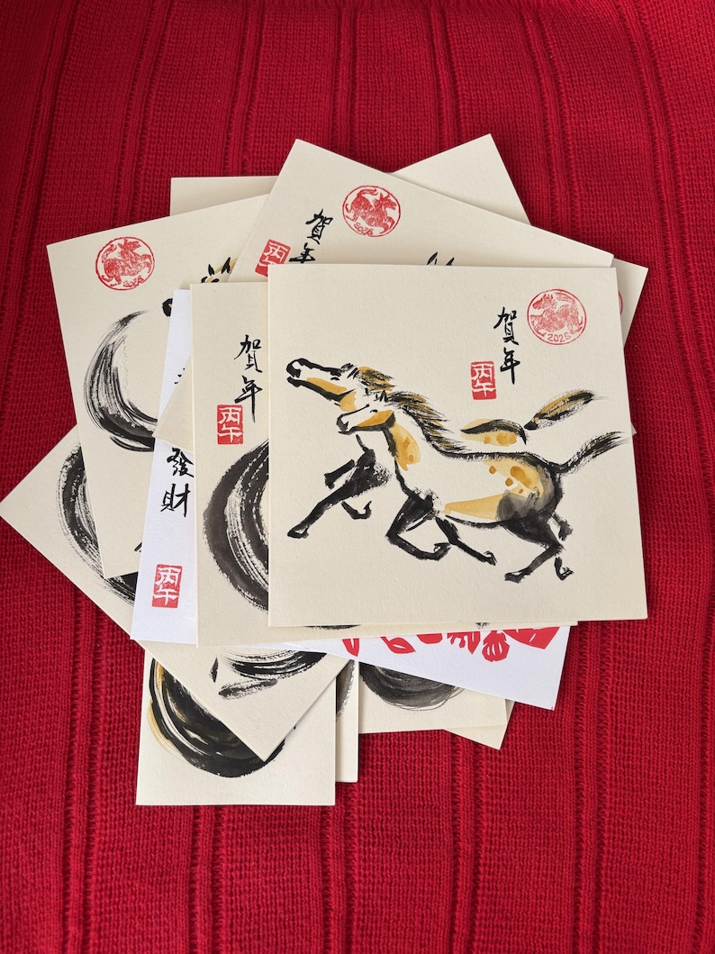 May include: A collection of square greeting cards with Japanese calligraphy and illustrations. The cards feature a running horse, brushstrokes, and red stamps. The background is a red textured surface.