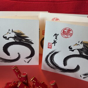 Pode incluir: Cart&otilde;es com uma ilustra&ccedil;&atilde;o de cavalo em tinta preta, com um carimbo circular vermelho e caracteres japoneses. Os cart&otilde;es est&atilde;o empilhados numa superf&iacute;cie vermelha, com objetos decorativos vermelhos em primeiro plano.
