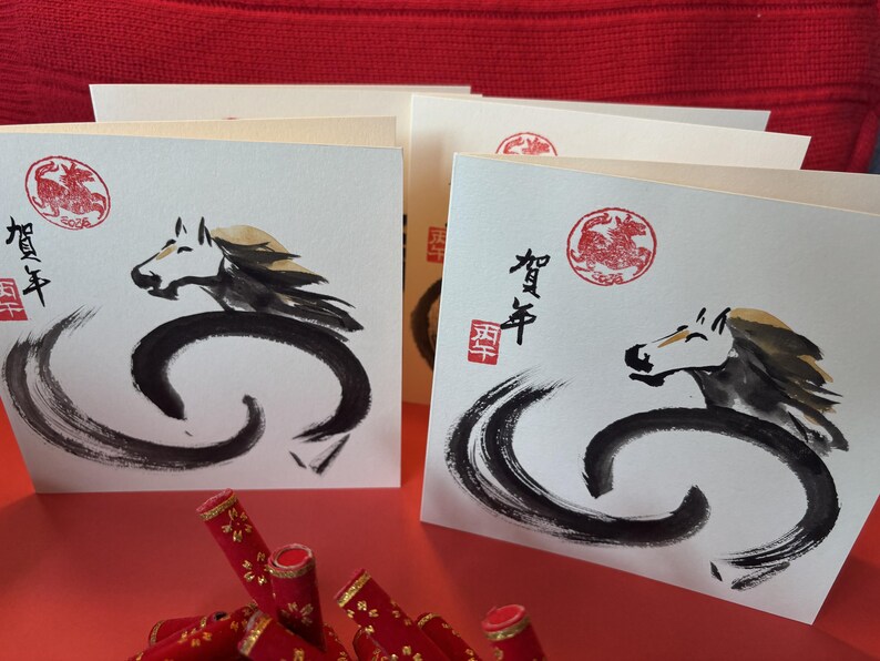 Pode incluir: Cart&otilde;es com uma ilustra&ccedil;&atilde;o de cavalo em tinta preta, com um carimbo circular vermelho e caracteres japoneses. Os cart&otilde;es est&atilde;o empilhados numa superf&iacute;cie vermelha, com objetos decorativos vermelhos em primeiro plano.