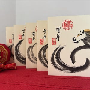 Pode incluir: Cart&otilde;es de felicita&ccedil;&otilde;es creme com caligrafia japonesa e ilustra&ccedil;&atilde;o de cavalo em tinta preta. Os cart&otilde;es est&atilde;o dispostos sobre uma superf&iacute;cie vermelha, com um objeto decorativo vermelho e dourado e petardos de papel vermelho em primeiro plano.