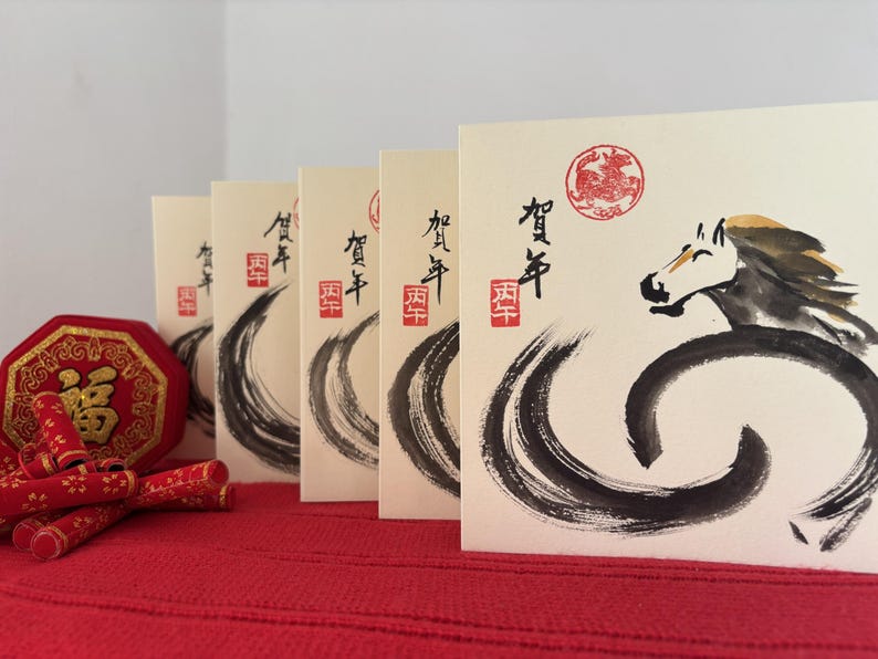 Pode incluir: Cart&otilde;es de felicita&ccedil;&otilde;es creme com caligrafia japonesa e ilustra&ccedil;&atilde;o de cavalo em tinta preta. Os cart&otilde;es est&atilde;o dispostos sobre uma superf&iacute;cie vermelha, com um objeto decorativo vermelho e dourado e petardos de papel vermelho em primeiro plano.