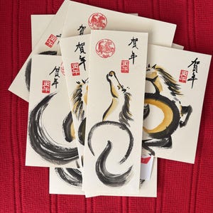 K&ouml;nnte beinhalten: Eine Sammlung cremefarbener Karten mit Tuschezeichnungen von Pferden in Schwarz und Gold. Jede Karte hat japanischen Text und einen roten runden Stempel. Die Karten sind auf einer roten strukturierten Oberfl&auml;che angeordnet.