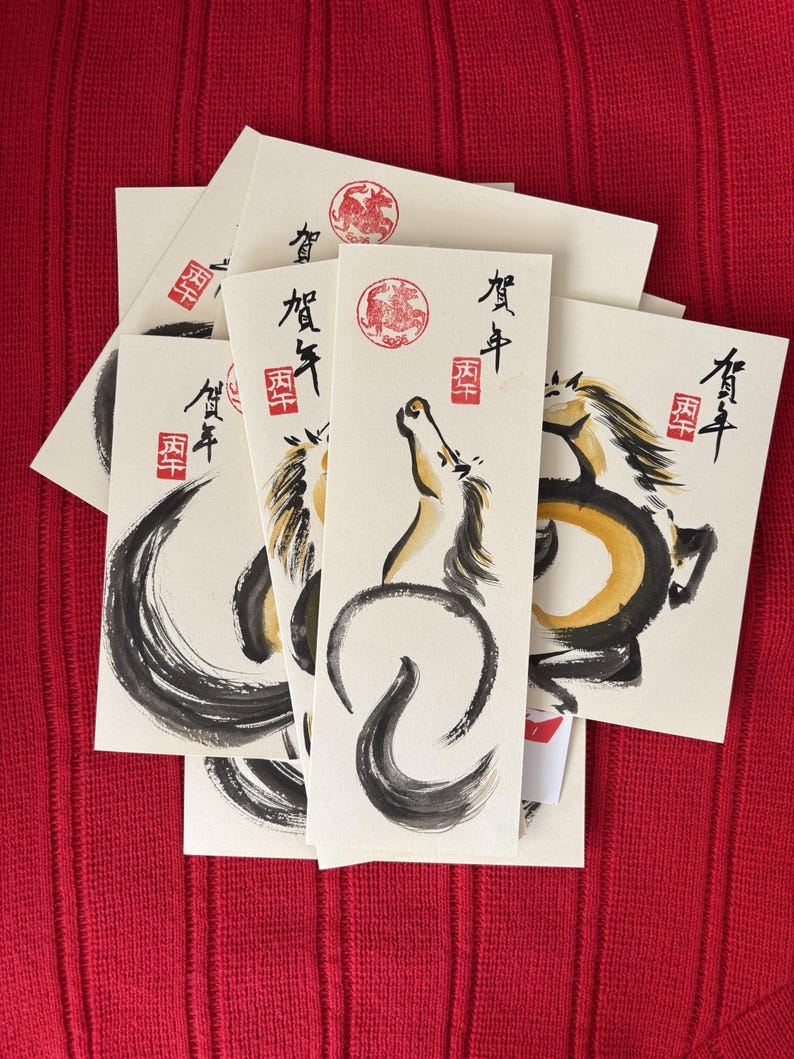 K&ouml;nnte beinhalten: Eine Sammlung cremefarbener Karten mit Tuschezeichnungen von Pferden in Schwarz und Gold. Jede Karte hat japanischen Text und einen roten runden Stempel. Die Karten sind auf einer roten strukturierten Oberfl&auml;che angeordnet.