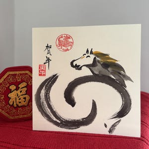 Pode incluir: Cart&atilde;o de felicita&ccedil;&otilde;es quadrado com uma pintura em aquarela de um cavalo em tinta preta e cinza. O cavalo tem uma crina amarela e est&aacute; sobre um fundo creme. Caracteres chineses e um selo vermelho no canto superior esquerdo.