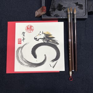 Pode incluir: Um conjunto de caligrafia com um cart&atilde;o de felicita&ccedil;&otilde;es com uma ilustra&ccedil;&atilde;o de cavalo e texto japon&ecirc;s, uma pedra de tinta preta, pinc&eacute;is e um carimbo vermelho. O cavalo &eacute; retratado em tinta preta e dourada.