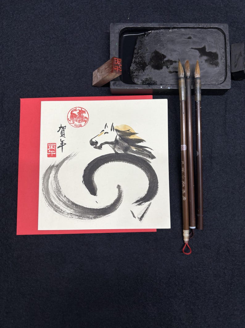 Pode incluir: Um conjunto de caligrafia com um cart&atilde;o de felicita&ccedil;&otilde;es com uma ilustra&ccedil;&atilde;o de cavalo e texto japon&ecirc;s, uma pedra de tinta preta, pinc&eacute;is e um carimbo vermelho. O cavalo &eacute; retratado em tinta preta e dourada.