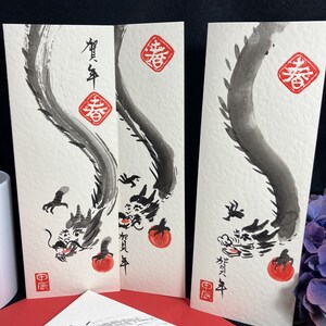 Puede incluir: Tres pinturas de dragones de tinta negra sobre papel blanco con caracteres chinos rojos y un sello rojo. Los caracteres en la pintura m&aacute;s a la izquierda dicen "A&ntilde;o Nuevo Chino". Los caracteres en la pintura del medio dicen "Primavera". Los caracteres en la pintura m&aacute;s a la derecha dicen "Primavera".
