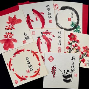 May include: A collection of Asian-inspired greeting cards featuring watercolor illustrations. The cards showcase red koi fish, flowers, and a panda, with calligraphy and red accents. The cards are on a black background.