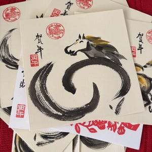 Pode incluir: Uma cole&ccedil;&atilde;o de cart&otilde;es quadrados com caligrafia japonesa e ilustra&ccedil;&otilde;es a tinta. Os cart&otilde;es apresentam um desenho de cavalo com pinceladas pretas e douradas, selos vermelhos e texto japon&ecirc;s. Os cart&otilde;es est&atilde;o empilhados numa superf&iacute;cie vermelha.