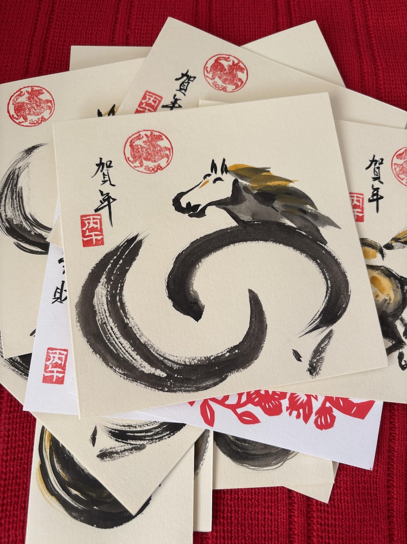 Pode incluir: Uma cole&ccedil;&atilde;o de cart&otilde;es quadrados com caligrafia japonesa e ilustra&ccedil;&otilde;es a tinta. Os cart&otilde;es apresentam um desenho de cavalo com pinceladas pretas e douradas, selos vermelhos e texto japon&ecirc;s. Os cart&otilde;es est&atilde;o empilhados numa superf&iacute;cie vermelha.