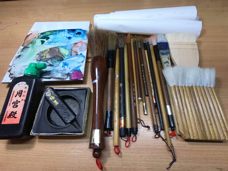 Może przedstawiać: Zestaw do kaligrafii, w skład kt&oacute;rego wchodzi butelka czarnego atramentu, kamień do atramentu, czarny pręt atramentowy, paleta akwarelowa i r&oacute;żne pędzle. Pędzle są wykonane z naturalnego włosia i mają drewniane uchwyty.