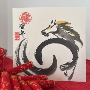 Pode incluir: Cart&atilde;o de felicita&ccedil;&otilde;es quadrado com uma ilustra&ccedil;&atilde;o de cavalo a tinta preta e dourada. O cart&atilde;o tem caracteres japoneses e um selo vermelho. Itens decorativos vermelhos com detalhes dourados est&atilde;o em primeiro plano.