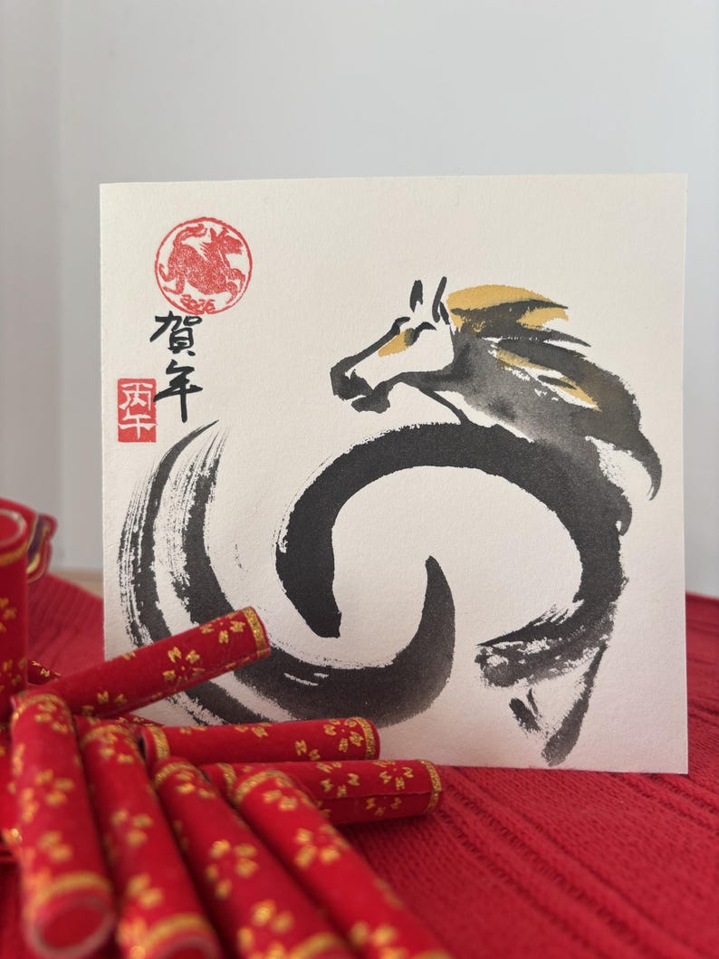Pode incluir: Cart&atilde;o de felicita&ccedil;&otilde;es quadrado com uma ilustra&ccedil;&atilde;o de cavalo a tinta preta e dourada. O cart&atilde;o tem caracteres japoneses e um selo vermelho. Itens decorativos vermelhos com detalhes dourados est&atilde;o em primeiro plano.