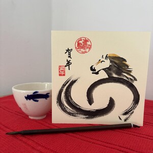 Pode incluir: Uma ch&aacute;vena de ch&aacute; de cer&acirc;mica branca com um desenho de peixe azul est&aacute; ao lado de um cart&atilde;o de felicita&ccedil;&otilde;es com uma ilustra&ccedil;&atilde;o de cavalo em tinta preta. O cart&atilde;o inclui texto japon&ecirc;s e um selo vermelho. Um pincel castanho escuro &eacute; colocado em frente ao cart&atilde;o.