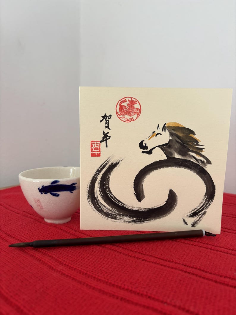 Pode incluir: Uma ch&aacute;vena de ch&aacute; de cer&acirc;mica branca com um desenho de peixe azul est&aacute; ao lado de um cart&atilde;o de felicita&ccedil;&otilde;es com uma ilustra&ccedil;&atilde;o de cavalo em tinta preta. O cart&atilde;o inclui texto japon&ecirc;s e um selo vermelho. Um pincel castanho escuro &eacute; colocado em frente ao cart&atilde;o.