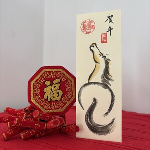 K&ouml;nnte beinhalten: Chinesische Neujahrsdekorationen in Rot und Gold. Enth&auml;lt eine achteckige rote Plakette mit goldenen Buchstaben, eine Karte mit einer Pferdeillustration und rote Feuerwerksk&ouml;rper-Ornamente. Der Hintergrund ist eine rote, strukturierte Oberfl&auml;che.
