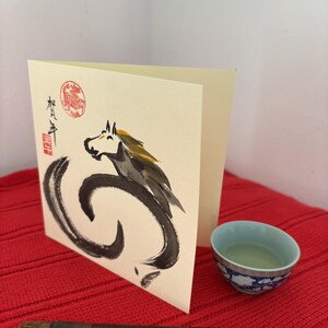Pode incluir: Um cart&atilde;o de felicita&ccedil;&otilde;es creme com uma ilustra&ccedil;&atilde;o de cavalo preto e cinza e caracteres japoneses. Uma pequena ch&aacute;vena de ch&aacute; azul e branca e um pincel numa superf&iacute;cie vermelha.